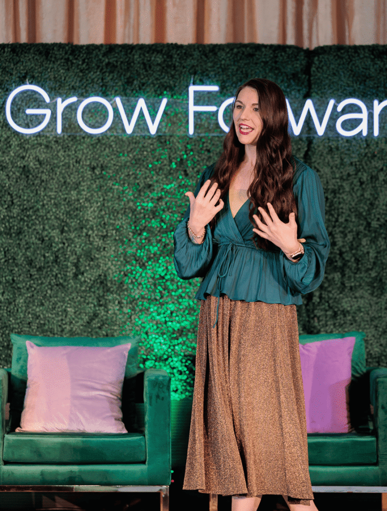 A woman speaks energetically on stage against a green backdrop with "Grow Forward." She's in a teal blouse and gold skirt, standing by green chairs with pillows.