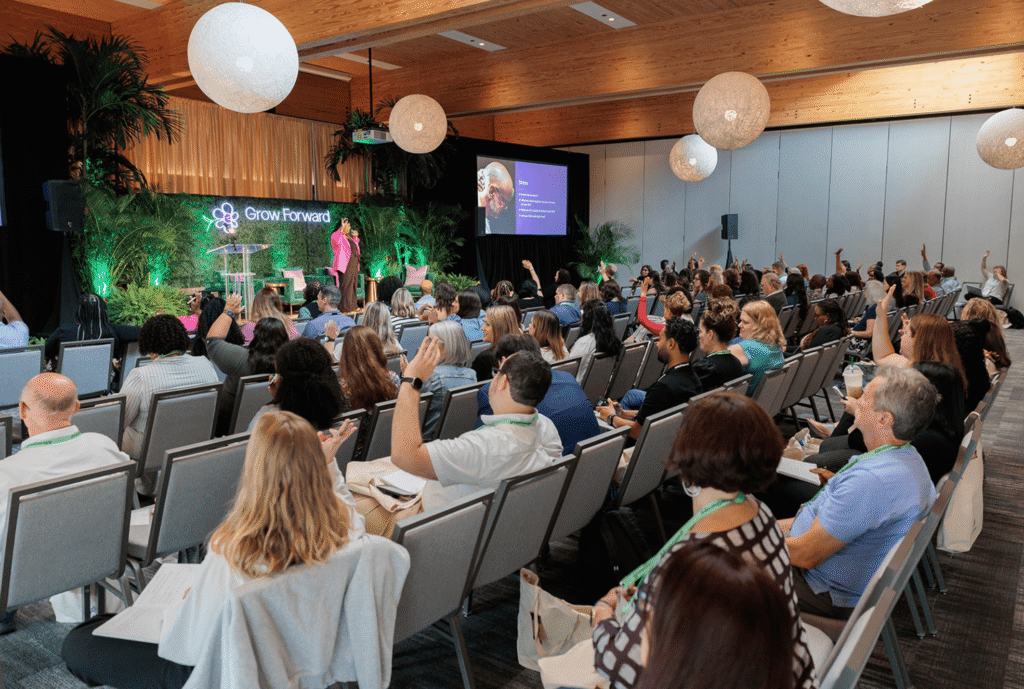 Audience seated in a conference room with a "Grow Forward" banner, listening to a speaker. Attendees raise hands, indicating engagement.