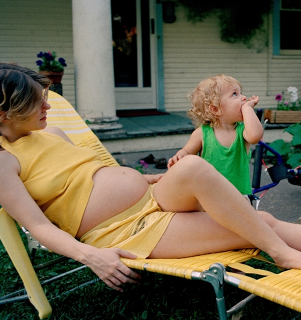 A pregnant woman in a yellow dress relaxes on a lounge chair outside, while a toddler beside her in a green shirt looks up thoughtfully, conveying warmth.