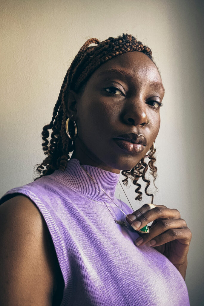 A confident woman in a purple top holds a necklace, her braided hair styled elegantly. She gazes forward with a calm expression, exuding poise.