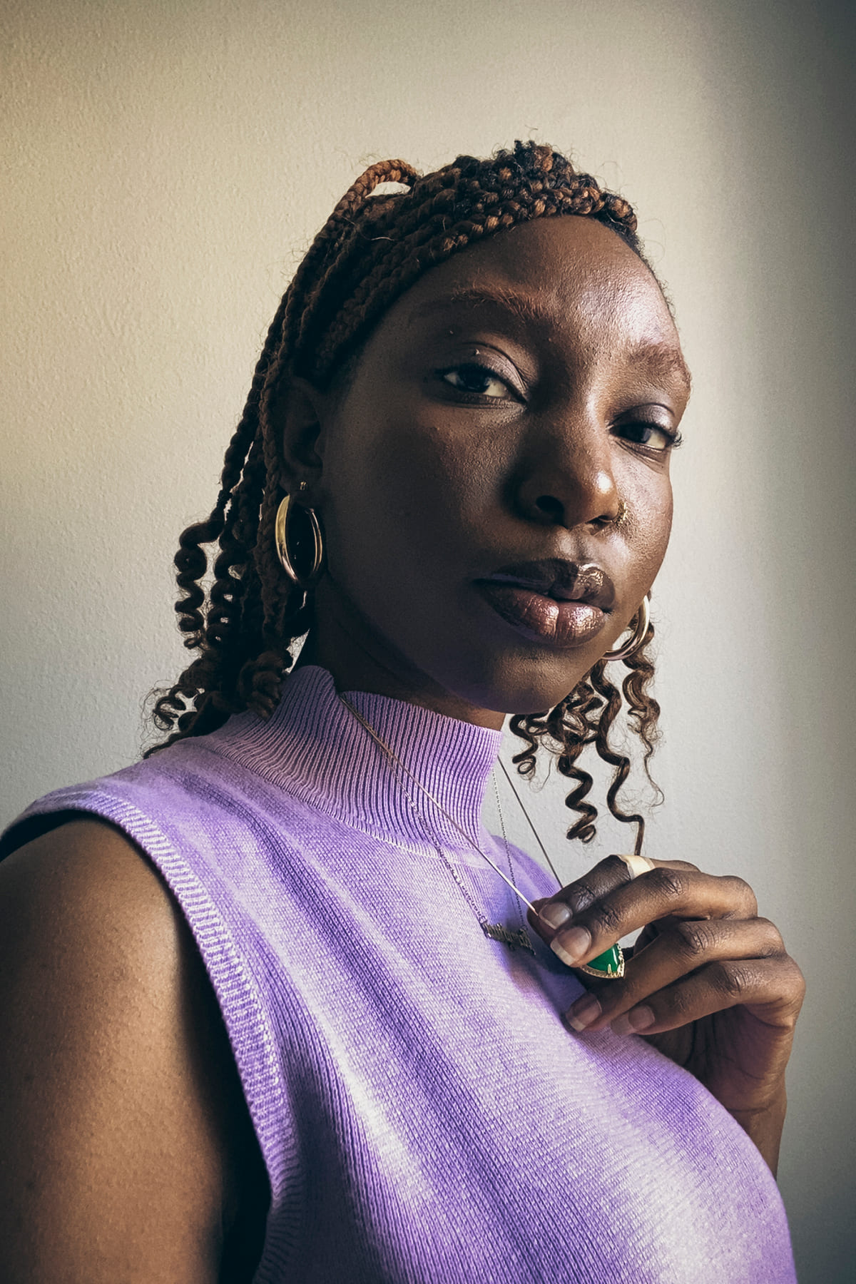 A confident woman in a purple top holds a necklace, her braided hair styled elegantly. She gazes forward with a calm expression, exuding poise.
